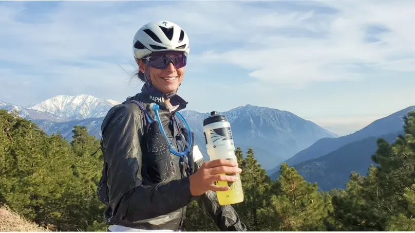 Michelle Howe, Professional trathlete, MS, CSSD, holding a bike bottle and wearing a jacket with a hydration pack, helmet, and sunglasses